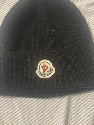 Svart Moncler mössa med logga - Säljer en svart ribbstickad mössa från Moncler med uppvikt kant och den klassiska Moncler-loggan framtill. Mössan är enkel men stilren och passar perfekt till kalla dagar. Materialet känns mjukt och bekvämt.