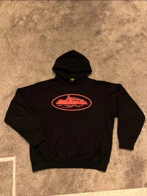 Svart Corteiz hoodie med röd grafik - Svart hoodie från Carhartt med stor röd grafisk print på bröstet i form av ett ovalt motiv. Klassisk känguruficka framtill och ribbade muddar vid ärmslut och nederkant. Tillverkad i mjukt bomullsmaterial och har en rymlig huva.