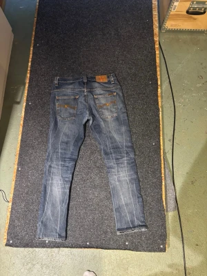 Blå slim/straight jeans från Nudie Jeans - Säljer ett par blå slim fit/ straight jeans från Nudie Jeans med klassiska fem fickor och snygga slitningar. Jeansen har kontrastsömmar, läderpatch bak i midjan och är tillverkade i bomull. Perfekta för dig som gillar en smalare passform och tidlös stil.