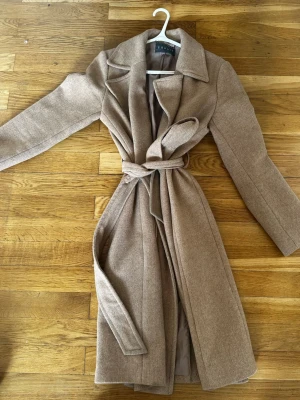 Beige kappa med knytskärp från  - Stilren beige kappa  med klassisk krage och knytskärp i midjan. Lång modell med raka ärmar och mjukt tyg som ger en snygg siluett. Perfekt för dig som gillar minimalistisk och trendig stil.