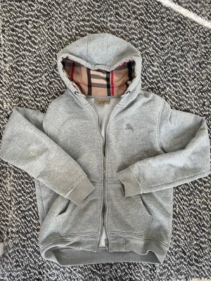 Grå hoodie från Burberry med dragkedja - Snygg grå hoodie från Burberry med klassiskt rutigt foder i huvan och insidan. Dragkedja framtill, två fickor och ribbade muddar. Perfekt för en stilren och avslappnad look. Ikonisk Burberry-logga broderad på bröstet.