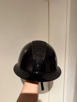 Svart ridhjälm med glitterdetalj - Helt ny hjälm från hansbo ridsport, aldrig använd då jag fick en annan. Har mips! Justerbar passform med ett ”hjul” på baksidan. Har även solskärm! Snygg också såklart😊