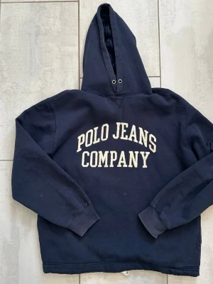 Mörkblå hoodie Polo Jeans Company - Snygg mörkblå hoodie från Polo Jeans Company med stor vit logga på bröstet. Boxy passform med huva och ribbade muddar vid ärmar och nederkant. Snöre insytt runt midjan för stt justera längd. Hål och fläck enligt bild på ena ärmen. Storlek M/L