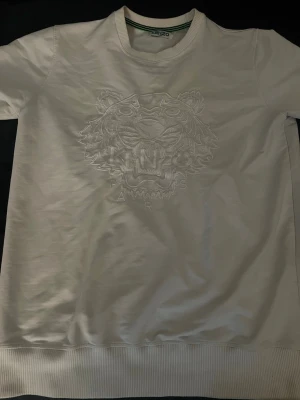 Vit sweatshirt med tiger - Säljer en vit sweatshirt med det ikoniska tigermotivet broderat på bröstet. Tröjan har rund halsringning, ribbade muddar och är tillverkad i mjukt bomullsmaterial. Perfekt för dig som gillar stilrena och trendiga plagg med exklusiv känsla.