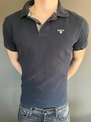 Mörkblå pikétröja från Barbour - Klassisk mörkblå pikétröja från Barbour med korta ärmar och krage. Tröjan har en liten logotyp på bröstet och rutigt tyg på insidan av kragen. Perfekt för en stilren och avslappnad look.