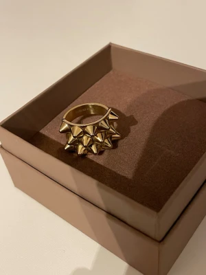 Guldig ring med nitar - Cool ring i guldfärgad metall med flera spetsiga nitar runt hela bandet. Ringen har en chunky och edgy design som verkligen sticker ut och passar dig som gillar statement-smycken. Perfekt för att ge din outfit en tuff touch. storlek 17.5