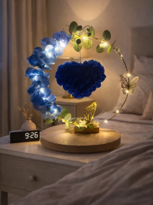 Dekorativ LED-lampa med blå rosor - Unik dekorationslampa med LED-slinga, blå konstgjorda rosor formade som ett hjärta, gröna blad och två guldfärgade fjärilar. Lampan står på en rund träbas och ger mysig belysning till rummet. Perfekt som inredningsdetalj eller present.