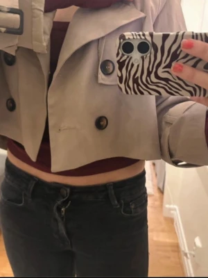 Kort beige trenchcoat med knappar - Säljer en trendig kort trenchcoat i beige med dubbelknäppning och bred krage. Jackan har stora svarta knappar och ett bälte i midjan. Perfekt för dig som vill ha en modern twist på klassikern. Materialet känns som bomull eller en bomullsblandning.