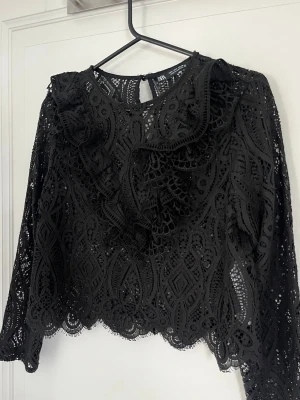 Svart spetsblus med volang från Zara - Supersnygg svart blus från Zara i spets med volangdetaljer framtill.🩷storlek S, inga defekter o använd fåtal ggr! 