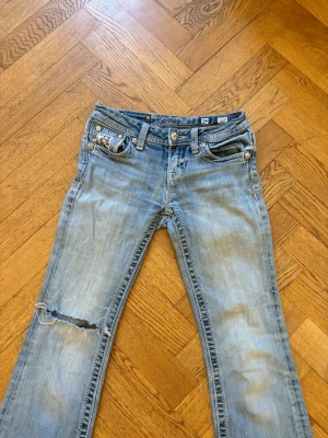 Miss me jeans  - Säljer ett par ljusblå bootcut jeans från miss me med snygga slitningar och hål på ena knät. Jeansen har låg midja och detaljerade nitar på fickorna. Perfekta för en avslappnad och trendig look. En knapp har lossnat på ena fickan och fickan är fastlimmad för annars sticker fickan ut väldigt mycket men inget man tänker på. 