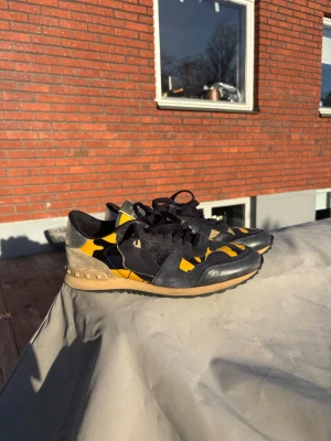 Valentino svart och gul sneaker - Säljer ett par snygga sneakers från Valentino med svart och gul camo-design. Skorna har svarta skosnören, beige hälparti och en klassisk låg siluett. Materialet är en mix av läder och textil, perfekt för dig som vill sticka ut med coola detaljer.