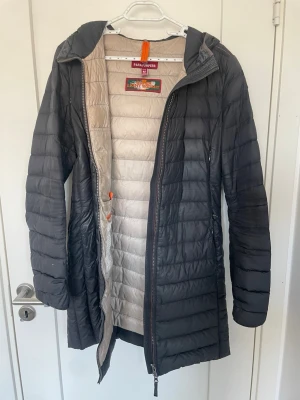 Svart lång dunjacka Parajumpers - Snygg svart lång dunjacka från Parajumpers med beige insida och huva. Jackan har quiltad design, dragkedja framtill och praktiska fickor. Perfekt för kalla vinterdagar och riktigt lätt att bära. Märket syns tydligt på insidan.