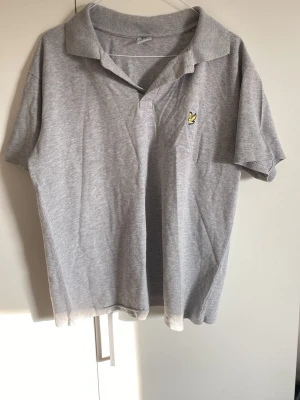 Grå pikétröja från Lyle & Scott - Klassisk grå pikétröja från Lyle & Scott med korta ärmar och gul logga på bröstet. Tröjan har krage och knäppning framtill. Perfekt till en avslappnad stil och enkel att matcha med jeans eller shorts. Storlek m men passar även s