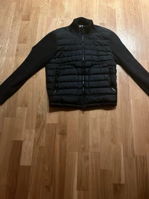 Svart cardiganjacka från Moncler - Snygg svart cardiganjacka från Moncler med quiltad front och stickade ärmar. Jackan har dragkedja framtill, två fickor med dragkedja och en liten Moncler-logga på ärmen. Perfekt för lager-på-lager och streetwear-vibe.