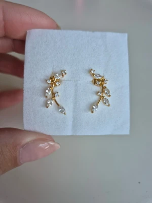 Örhängen, 18k guldpläterat äkta silver - Örhängen i 18k guldpläterat äkta silver med böjda bladmönster och dekorativa Cubic Zirconia stenar. Från Glitter, slutsålda på deras hemsida. Köpta som en julklapp som inte passade. Kvitto finns men tyvärr finns inte den 'brickan' dem ska sitta på kvar. Oanvända, nya.  Mått: 2 x 0,5 cm Material: 18k guldpläterat Sterling Silver 925 | Cubic Zirconia