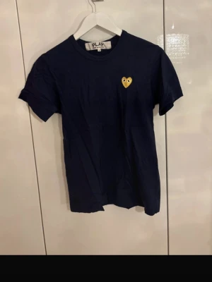 Mörkblå Comme des Garçons PLAY t-shirt - Snygg mörkblå t-shirt från Comme des Garçons PLAY med det ikoniska hjärta-märket i gult på bröstet. Klassisk passform och rund halsringning. Perfekt till jeans eller shorts för en clean och stilren look.