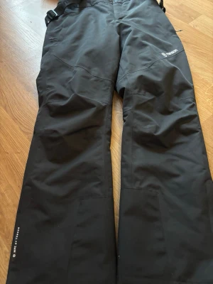 Svarta täckbyxor från Tenson - Säljer ett par svarta täckbyxor från Tenson med justerbara hängslen och förstärkta knän. Byxorna har två knappar i midjan och dragkedja, samt logga på benet. Perfekta för kalla dagar i backen eller vinteräventyr.