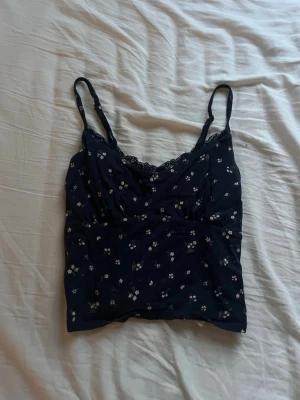 Mörkblå blommig linnetopp med spets - Söt mörkblå linnetopp med små vita blommor och tunna axelband. Toppen har spetsdetalj vid urringningen och är croppad i modellen. Perfekt för varma dagar eller som layering-piece. Den är i storleken S men passar mer som XS.