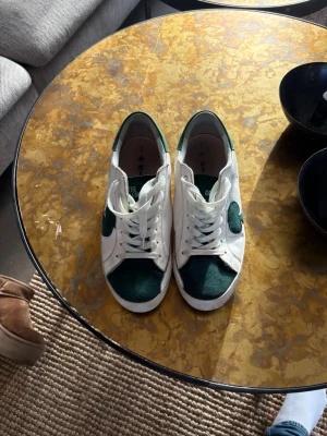 Gröna och vita sneakers från the gallerist  - Snygga sneakers från the gallerist i vitt och mörkgrönt. Skorna har lågt skaft, klassisk snörning och en kontrasterande grön detalj på sidan samt tåpartiet. Ovandelen är i mocka och textil, vilket ger en stilren och sportig look. Finns ej ute på hemsidan! Köpta för 2600
