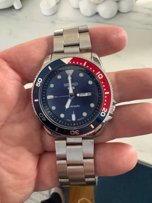 Seiko mod klocka blå/röd - Snygg Seiko mod  klocka med blå urtavla och vridring i blått och rött. Klockan har tydliga vita index, dag- och datumvisning samt ett robust armband i silverfärgad metall. Perfekt för dig som gillar sportig och klassisk stil. Riktigt blank och fin! Helt ny! 