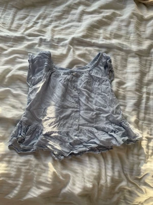 Blårandig blus med volangkant - Söt blårandig blus med korta ärmar och volangkant nedtill. Toppen har rund halsringning och knappar framtill. Perfekt för sommarens alla dagar och ger en avslappnad men ändå trendig look.