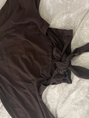 Brun omlottklänning med knytband - Snygg brun omlottklänning med breda knytband i midjan som ger en fin siluett. Klänningen har en enkel, elegant design och är tillverkad i ett mjukt tyg som känns skönt mot huden. Perfekt för dig som gillar stilrena plagg med det lilla extra.
