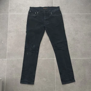 Svarta jeans från Nudie Jeans - Säljer ett par svarta jeans från Nudie Jeans med klassisk femficksdesign och diskret logga på bakfickan. Jeansen har smal passform och raka ben, tillverkade i mjukt denimtyg. Perfekta för dig som gillar stilrena och tidlösa jeans. W32 L32. Modell: GrimTim