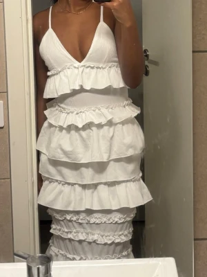 Vit långklänning med volanger - Superfin vit långklänning med smala axelband och djup v-ringning. Klänningen har flera lager volanger som ger en riktigt snygg och trendig look. Perfekt för sommarens alla tillfällen och ger en romantisk vibe.