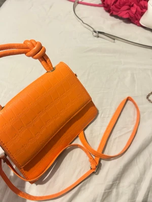 Orange croco-inspirerad handväska - Snygg orange handväska i croco-mönstrat konstläder med axelrem och coolt knutdetaljerat handtag. Väskan har en stilren, rektangulär form och stängs med lock. Perfekt för att addera färg till din outfit.