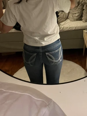 Mörkblå bootcut jeans med vit söm - Snygga mörkblå bootcut jeans med markerade vita sömmar och dubbla knappar fram. Bakfickorna har ett unikt broderat mönster i vitt som ger extra edge. Jeansen har låg midja och klassisk femficksdesign. Perfekta för dig som gillar en trendig och cool stil.