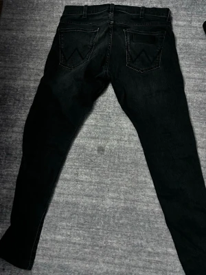 Svarta jeans från Wrangler Bryson - Säljer ett par svarta jeans från Wrangler, modell Bryson. Jeansen har klassisk femficksdesign, raka ben och tydliga sömmar på bakfickorna. Tillverkade i slitstarkt denimtyg som ger en snygg och tidlös look.