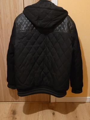 Svart quiltad pufferjacka från Smog - Snygg svart pufferjacka från Smog med quiltat mönster, huva och detaljer i fuskläder på axlarna. Jackan har dragkedja framtill, två bröstfickor med dragkedja och ribbade muddar vid ärmslut och nederkant. Perfekt för kalla vinterdagar.
