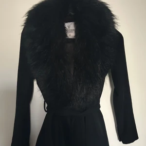 Svart kappa med fuskpäls från Chiara Forthi - Stilren svart kappa från Chiara Forthi med bred krage i svart fuskpäls. Pälsen är avtagbar. Kappan har knytband i midjan för en snygg siluett och är helsvart i färgen. Mycket bra skick, det enda är två öglor som till knapparna på pälsen är av, se bilder. 