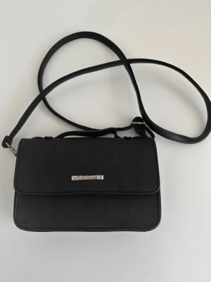 Svart väska från Björn Borg - Stilren svart axelväska / handväska / väska från Björn Borg i strukturerat konstläder. Väskan har ett avlångt lock, justerbar axelrem och en diskret guldfärgad logga framtill. Perfekt för att bära dina viktigaste grejer med snygg och enkel design. Har några bläckstänk i tyget inne på botten som ej gått bort vid rengöring, se bild, men inget som syns när det är saker i. Annars i mycket gott skick, som ny. 