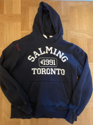 Marinblå hoodie Salming Toronto - Mörkblå hoodie från Salming med vit text 'Salming 1991 Toronto' på bröstet. Klassisk känguruficka framtill och huva. Perfekt för en sportig och avslappnad stil. Materialet är mjukt och bekvämt, passar till chill eller träning.