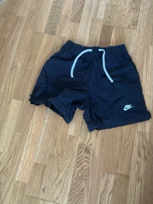 Mörkblå Nike shorts med snörning - Säljer ett par mörkblå shorts från Nike med vit snörning i midjan och logga på ena benet. Shortsen har en avslappnad passform och elastisk midja, perfekta för sommardagar eller träning.