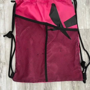 Rosa gympåse med svart meshficka - Säljer en rosa gympåse med svarta detaljer och snören. Framsidan har en stor meshficka och en svart tryckt detalj. Baksidan har en dragkedjeficka för extra förvaring. Perfekt för träning eller skola, lätt att bära på ryggen.