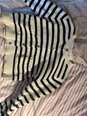 Randig stickad kofta med knappar - Säljer en svartvit stickad kofta med horisontella ränder och rund hals. Koftan har knäppning framtill med fem knappar och två små fickor på magen. Materialet är mjukt och koftan har en croppad passform som ger en trendig look.