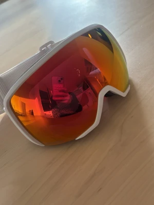 Vita HD sportiga solglasögon - Säljer ett par vita skidglasögon från Atomic med bred elastisk rem och röd-orange spegellins. Glasögonen har mjuk skumvaddering i svart och rött för extra komfort. Helt nya kommer med den originella lådan och tillbehören. Orginella priset var 899 tror jag.