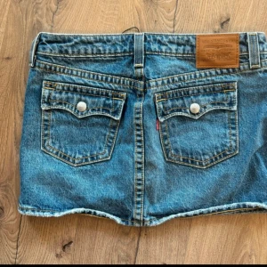 Blå jeanskjol från Levi's mini skirt  - Säljer en klassisk blå jeanskjol från Levi's med fem fickor, bälteshällor och knappdetaljer. Kjolen har en kort passform och är tillverkad i slitstark denim med en rå kant nertill. Perfekt för dig som gillar en avslappnad och trendig stil.