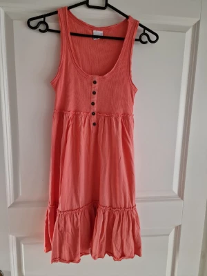 Korallrosa ärmlös sommarklänning  - Söt ärmlös klänning i korallrosa bomull med knappar framtill och volang nertill. Perfekt för varma dagar med sin luftiga och lediga passform. Rund halsringning och utan mönster för en enkel och fräsch look.