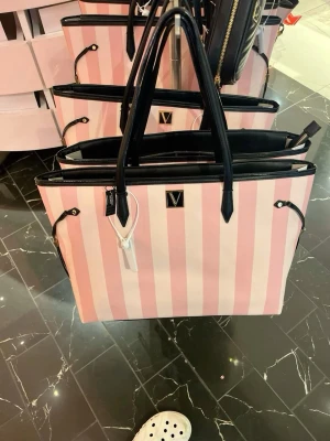 Tote väska - Snygg shoppingväska från Victoria's Secret med rosa och vita vertikala ränder. Väskan har svarta handtag och detaljer samt ett guldfärgat V-emblem framtill. Tillverkad i slitstarkt tygmaterial och har rymlig insida för allt du behöver. Bara en liten defekt som syns på sista bilden!