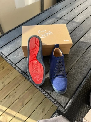 Blåa Christian Louboutin sneakers - Snygga blåa sneakers från Christian Louboutin med klassisk röd sula och ovandel i mocka. Skorna har rund tå, matchande blå skosnören och diskreta sömdetaljer. Perfekta för dig som vill sticka ut med exklusiv stil och ikonisk design.