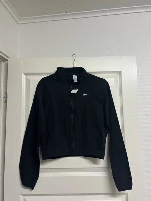 Svart Alo jacka – helt ny - Säljer en svart jacka från Alo Yoga. Helt ny och aldrig använd, lappen sitter kvar. Sportig och stilren cropped modell med dragkedja framtill. Lätt material som passar perfekt till både träning och vardag.  Mycket snygg passform och enkel att matcha med träningskläder eller jeans.