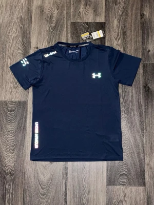 Mörkblå sport-tshirt från Under Armour - Mörkblå sport-tshirt från Under Armour med reflekterande logga och detaljer på bröstet, axeln och ärmen. Tillverkad i ett lätt och snabbtorkande funktionsmaterial som passar perfekt till träning. Rund halsringning och korta ärmar.