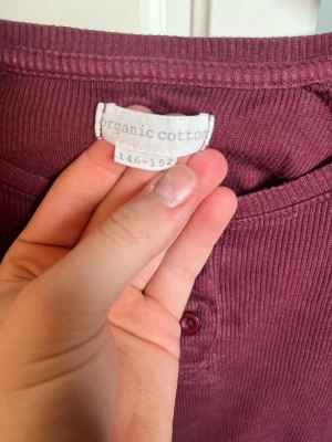 Vinröd ribbad långärmad tröja - Vinröd långärmad tröja i ribbad ekologisk bomull. Tröjan har rund halsringning och knappar framtill. Perfekt för dig som gillar en enkel och bekväm stil.