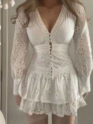 Den super populära fashion nova klänningen!  - Supersöt vit kort klänning med v-ringning, spetsdetaljer och volanger. Klänningen har långa ärmar med broderade blommor och knappar framtill. Perfekt för sommarens alla tillfällen och ger en romantisk vibe.