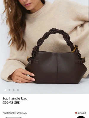 Mörkbrun handväska med flätat handtag - Snygg mörkbrun handväska i konstläder med unikt flätat handtag och guldfärgade metalldetaljer. Väskan har en mjuk, rundad form och justerbar axelrem för extra flexibilitet. Perfekt accessoar för att lyfta din outfit. Använd 1 gång