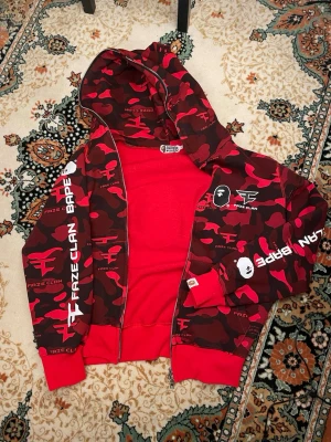 BAPE x FaZe Clan Zip Hoodie - Snygg röd camo hoodie från FaZe Clan x BAPE med dragkedja och stor huva. Hoodien har vita logotyper och text på ärmar och bröst, samt detaljer i svart och vitt. Tillverkad i mjukt bomullsmaterial och har ribbade muddar vid ärmslut och nederkant. Storlek L men sitter som S/XS