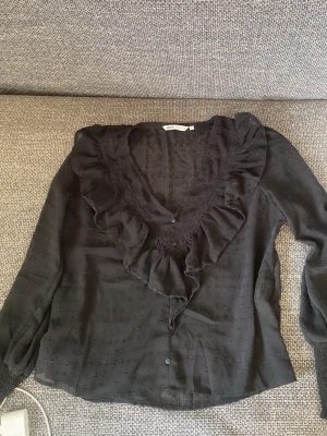 Svart blus med volang från Only  - Snygg svart blus från Only  med volangdetaljer längs den djupa v-ringningen. Blusen är i ett tunt, transparent tyg med diskret strukturmönster och har långa ärmar. Perfekt att styla med jeans eller kjol för en trendig look.